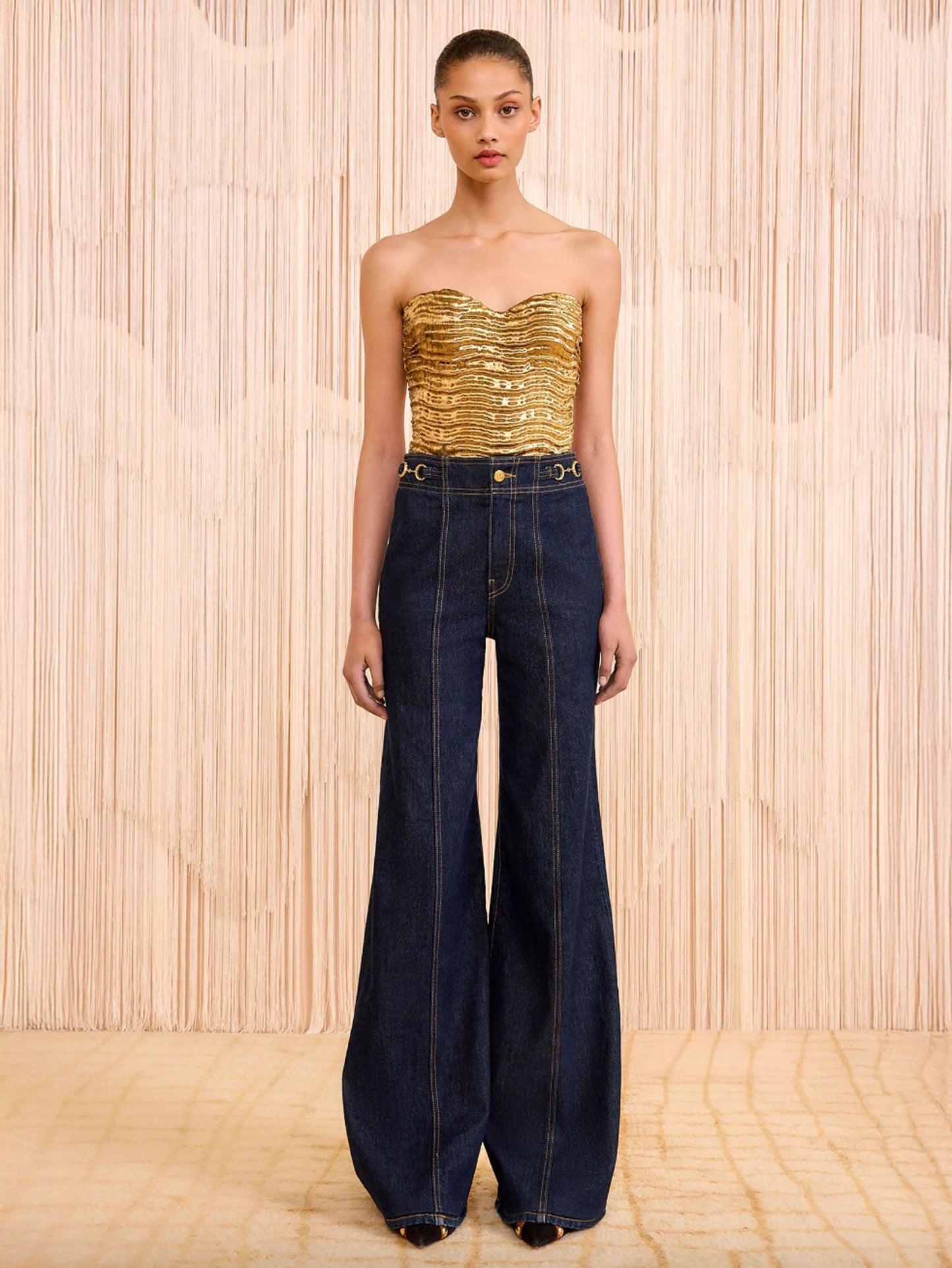 Model wearing a gold strapless top and blue jeans against a wooden background