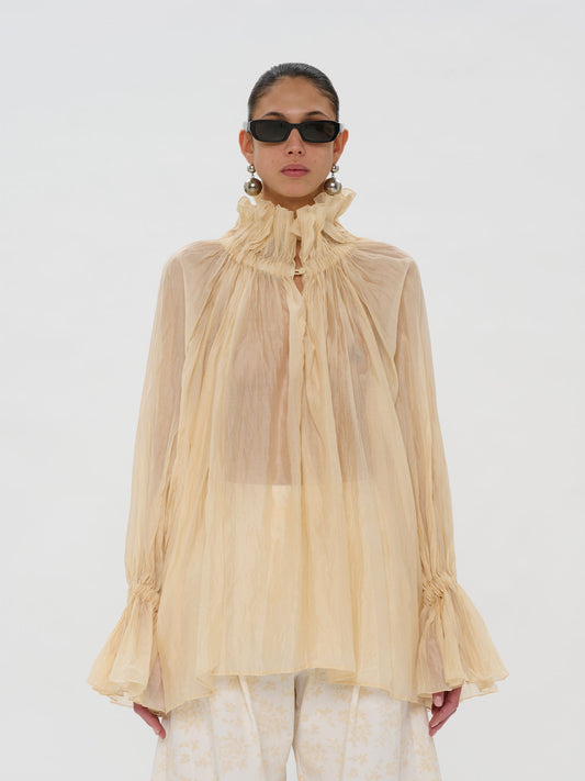 A model wearing a sheer, high-neck beige blouse with dramatic ruffles and long bell sleeves, paired with light patterned trousers and black sunglasses, against a plain white studio background.