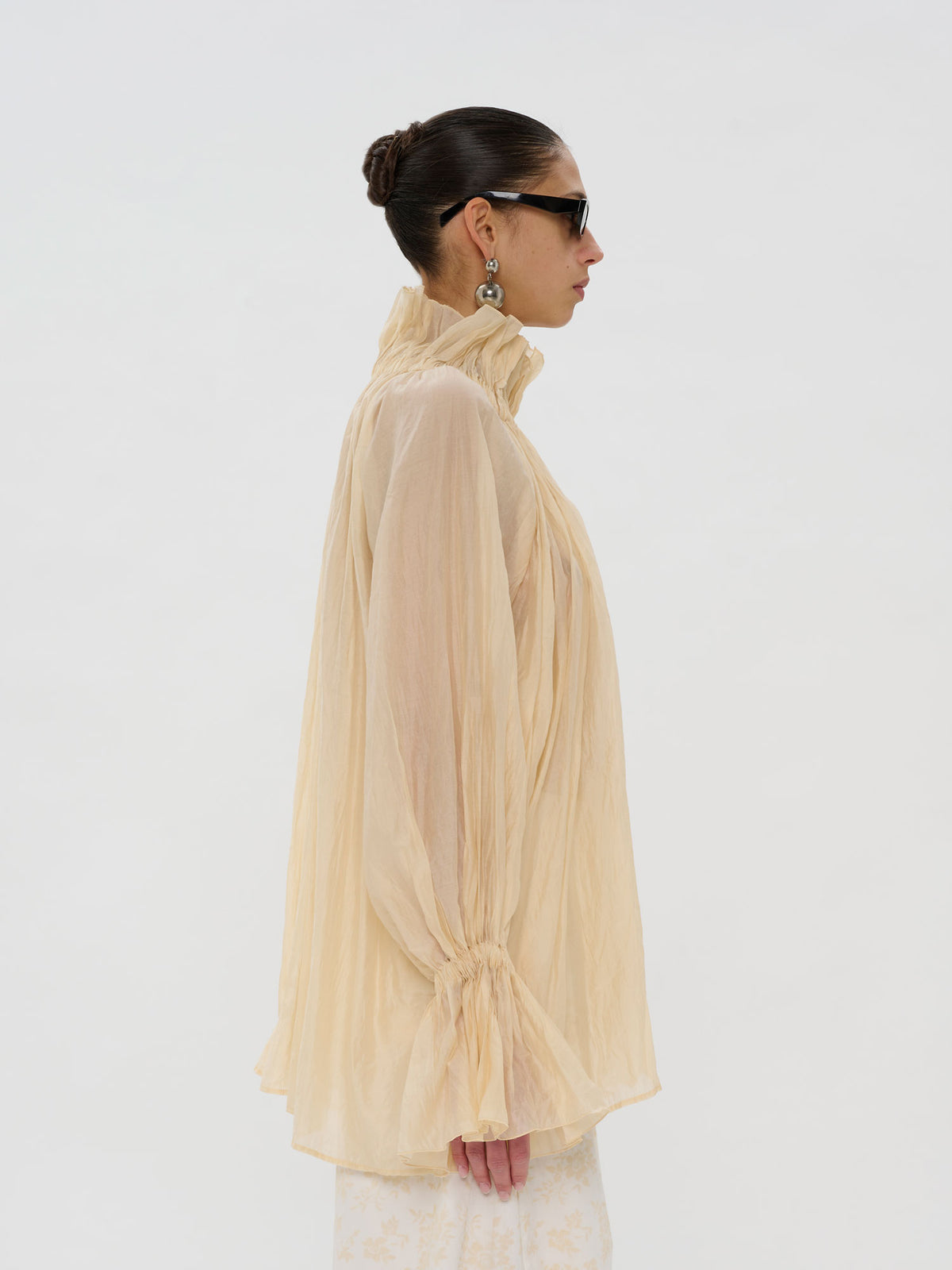 A model shown in profile wearing a sheer, high-neck, long-sleeve blouse in a pale peach-beige color, styled with black sunglasses and earrings against a plain white studio background.