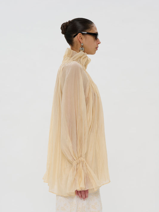 A model shown in profile wearing a sheer, high-neck, long-sleeve blouse in a pale peach-beige color, styled with black sunglasses and earrings against a plain white studio background.