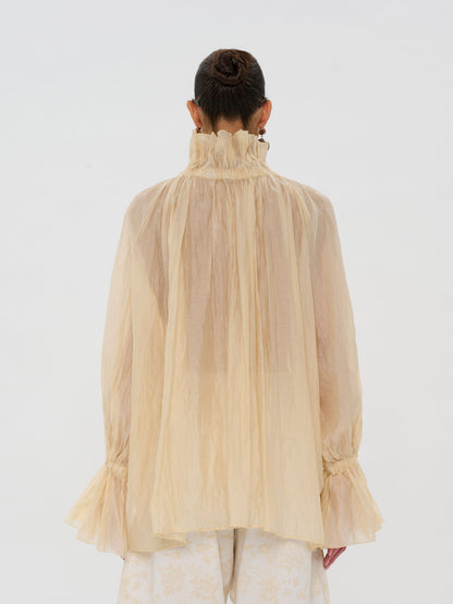 A model shown from the back wearing a sheer, high-neck beige blouse with long ruffled sleeves and gathered cuffs, paired with light patterned trousers, against a plain white studio background.