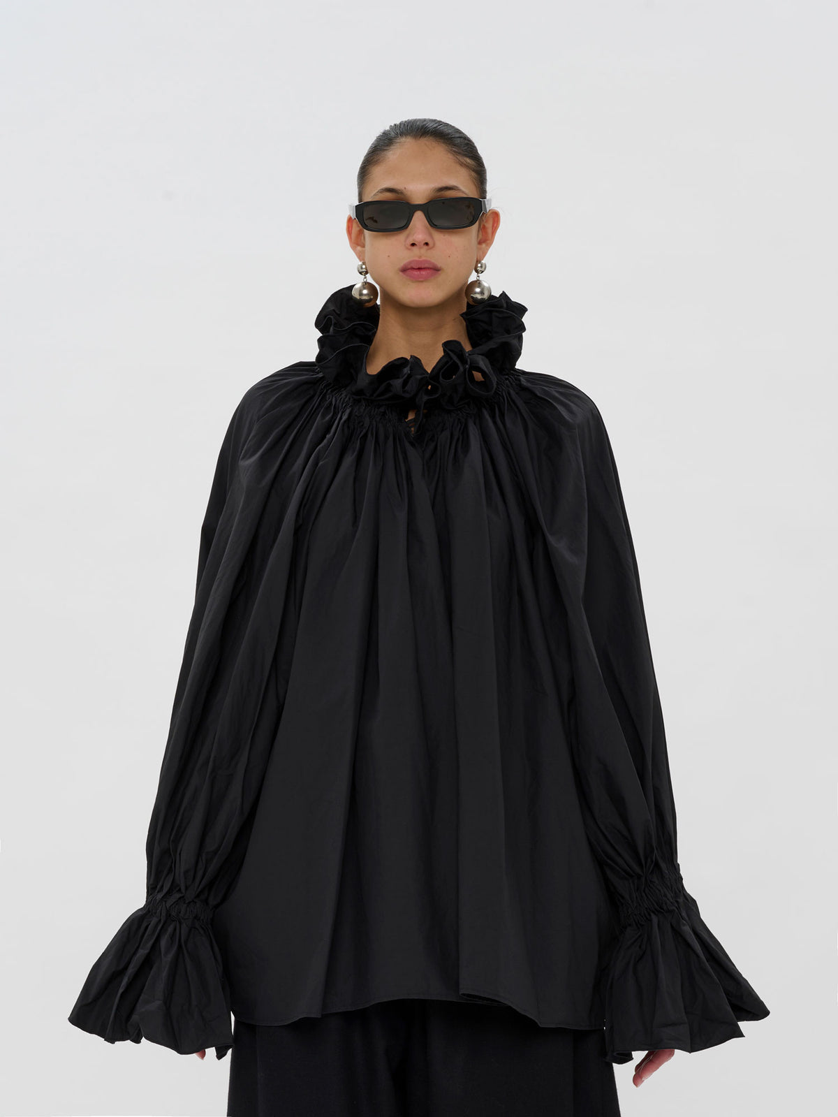 A model wearing a black long-sleeve blouse with a dramatic ruffled high collar, gathered sleeves, and flared cuffs, paired with black pants and sunglasses, against a plain light gray background.