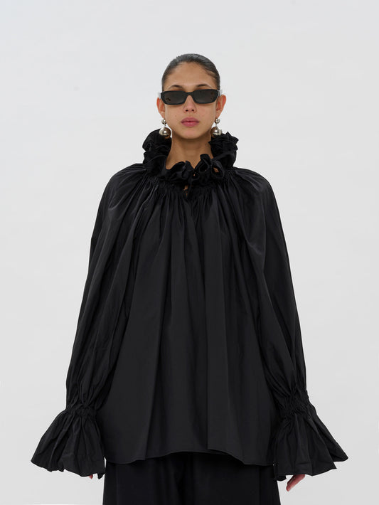 A model wearing a black long-sleeve blouse with a dramatic ruffled high collar, gathered sleeves, and flared cuffs, paired with black pants and sunglasses, against a plain light gray background.