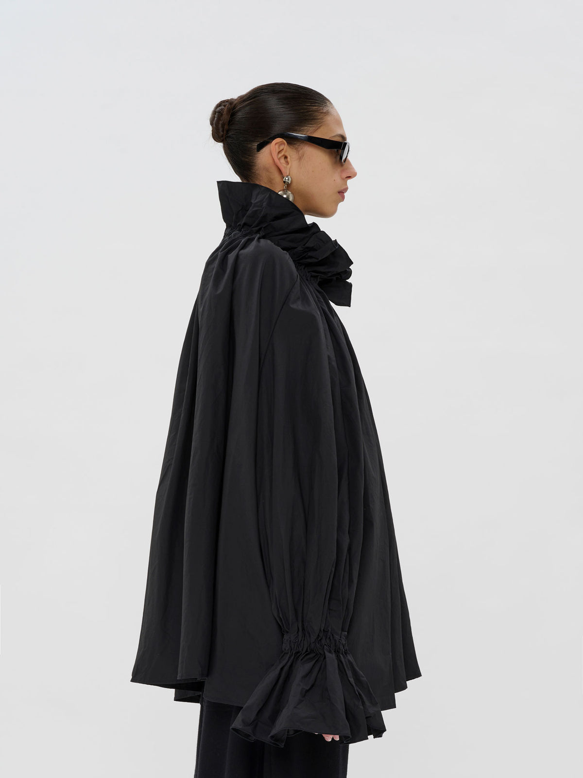 A model in profile wearing a black oversized cape-style jacket with a high ruffled collar and gathered long sleeves, photographed against a plain light gray studio background.