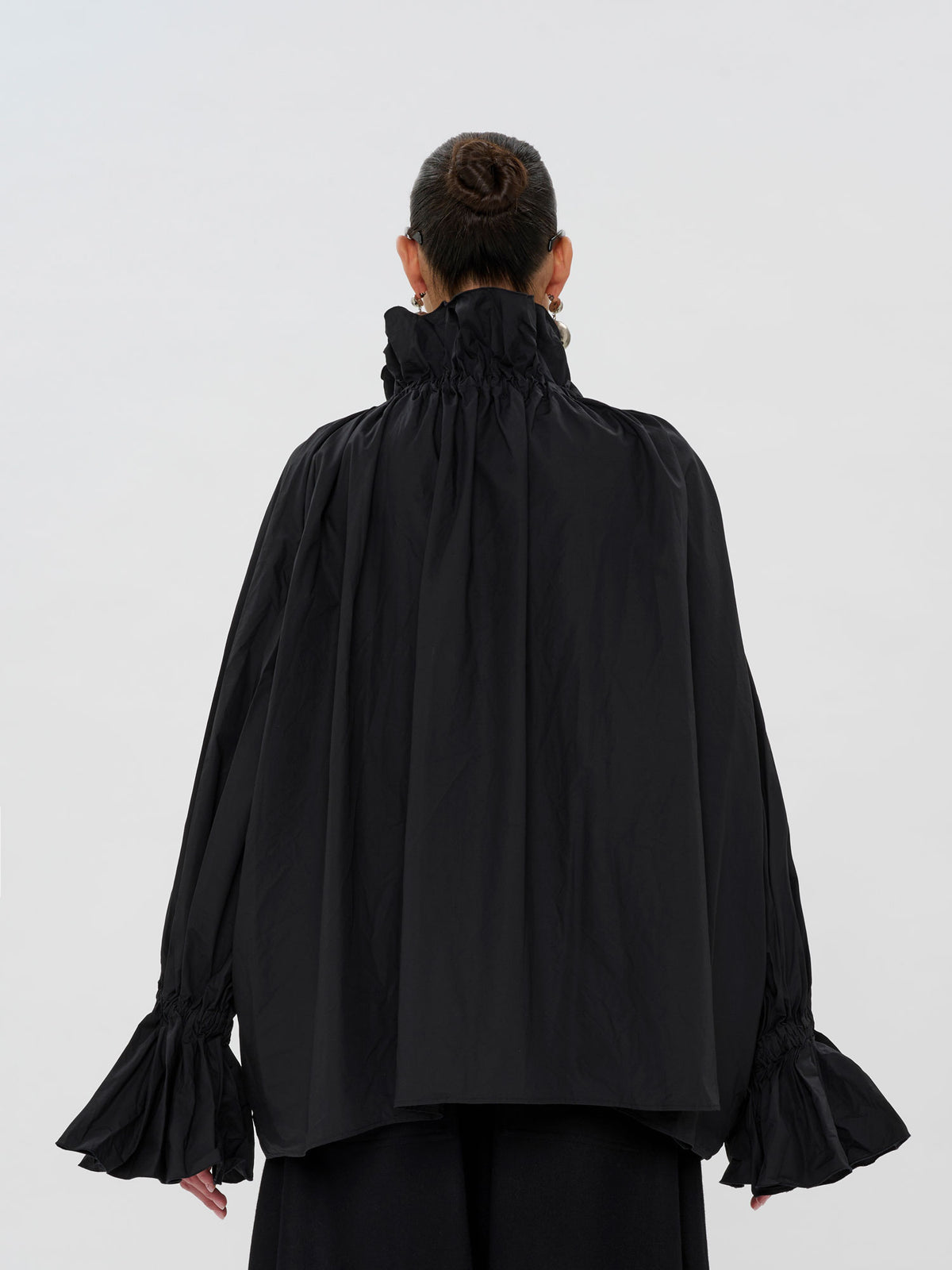 A model viewed from the back wearing a black long-sleeve blouse with a high gathered ruffled collar and flared bell sleeves, paired with black trousers, against a light gray studio background.