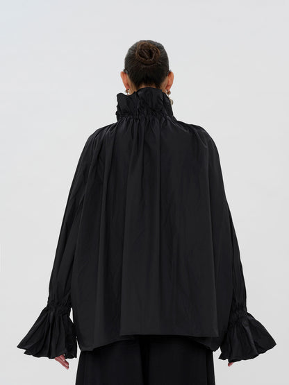A model viewed from the back wearing a black long-sleeve blouse with a high gathered ruffled collar and flared bell sleeves, paired with black trousers, against a light gray studio background.