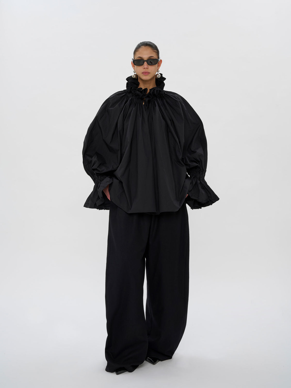 A model wearing a black oversized high-neck top with dramatic gathered sleeves and matching black wide-leg trousers, photographed against a seamless light gray studio background.