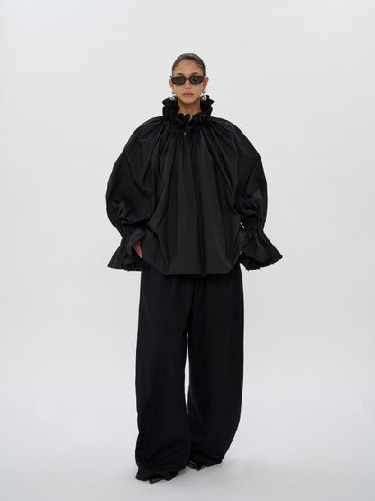 A model wearing a black oversized high-neck top with dramatic gathered sleeves and matching black wide-leg trousers, photographed against a seamless light gray studio background.