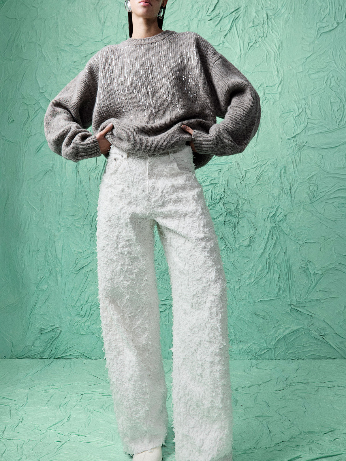 A model wearing a gray knit sweater with sequins and textured white wide-leg pants, standing against a textured mint green backdrop.