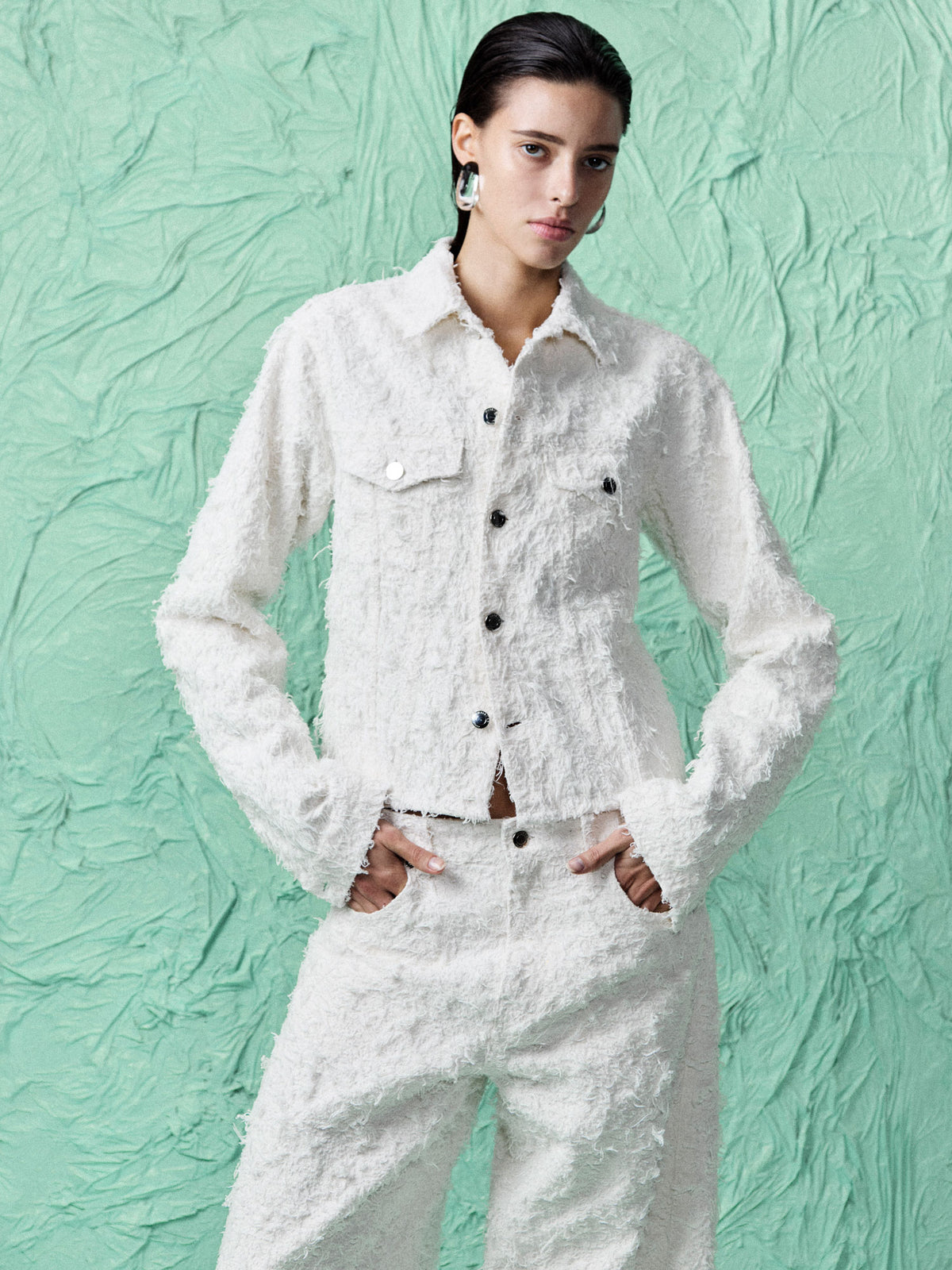 A model wearing a textured white long-sleeve button-up shirt and matching textured white trousers, standing against a textured mint green background.