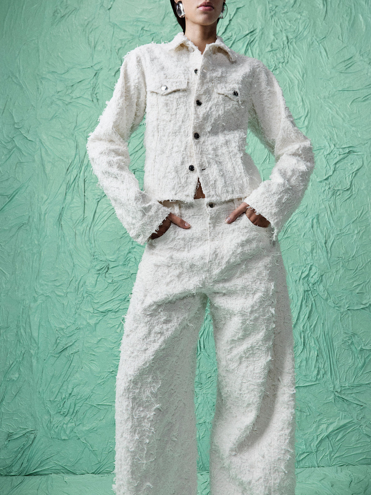 A model wearing a textured white tweed jacket and matching wide-leg trousers, standing against a textured mint green backdrop.