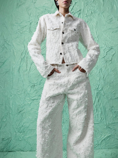 A model wearing a textured white tweed jacket and matching wide-leg trousers, standing against a textured mint green backdrop.