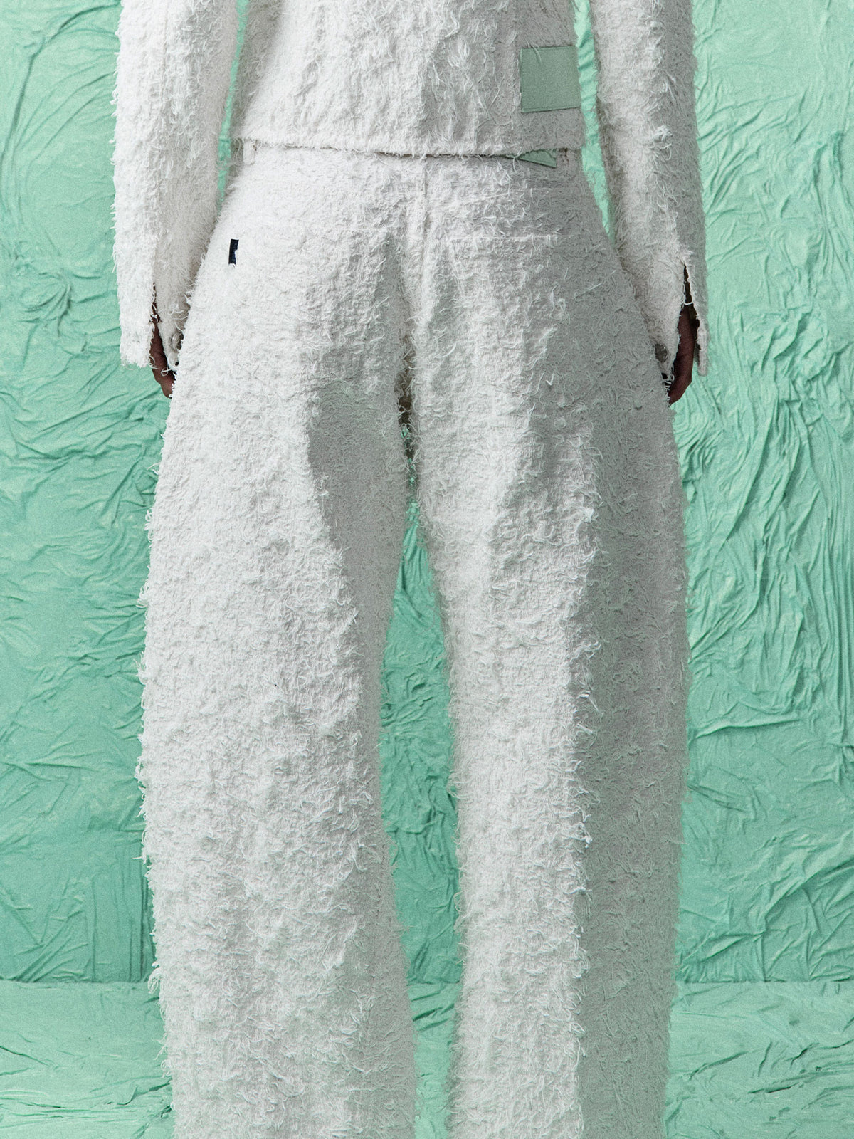 White textured bouclé pants with a matching cropped top, shown from the back on a model against a textured mint green backdrop.