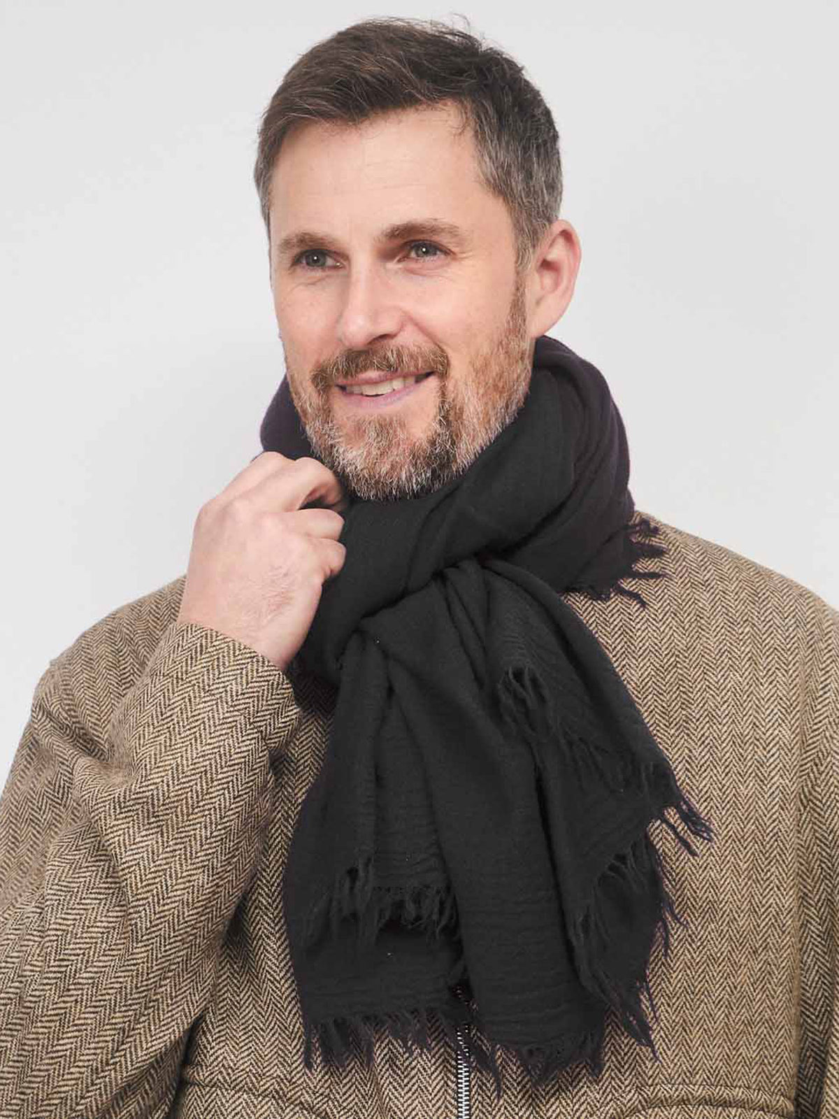 A man wearing a black fringed scarf and a brown herringbone jacket, posing against a light gray background.