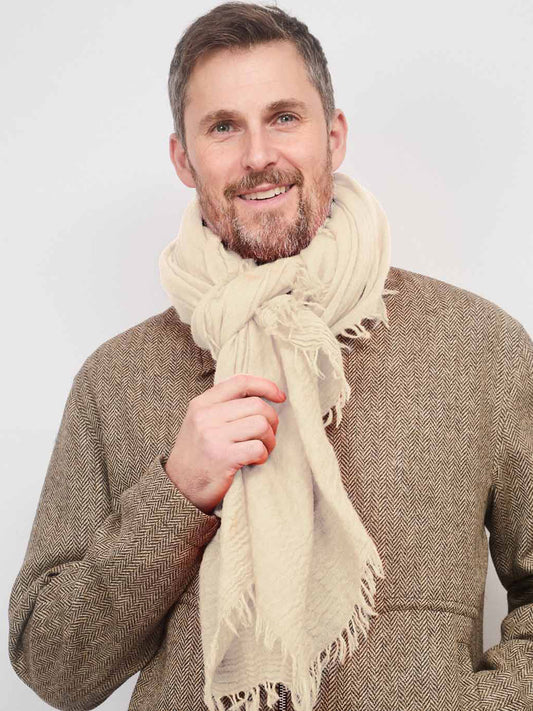 A smiling man wearing a thick cream-colored knit scarf with fringed edges over a brown herringbone tweed sweater, set against a plain light gray background.