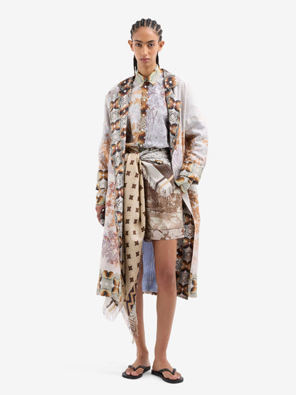 A model stands against a light gray studio background wearing a long, loose printed coat over a matching patterned shirt and shorts, with a patterned belt and sandals.