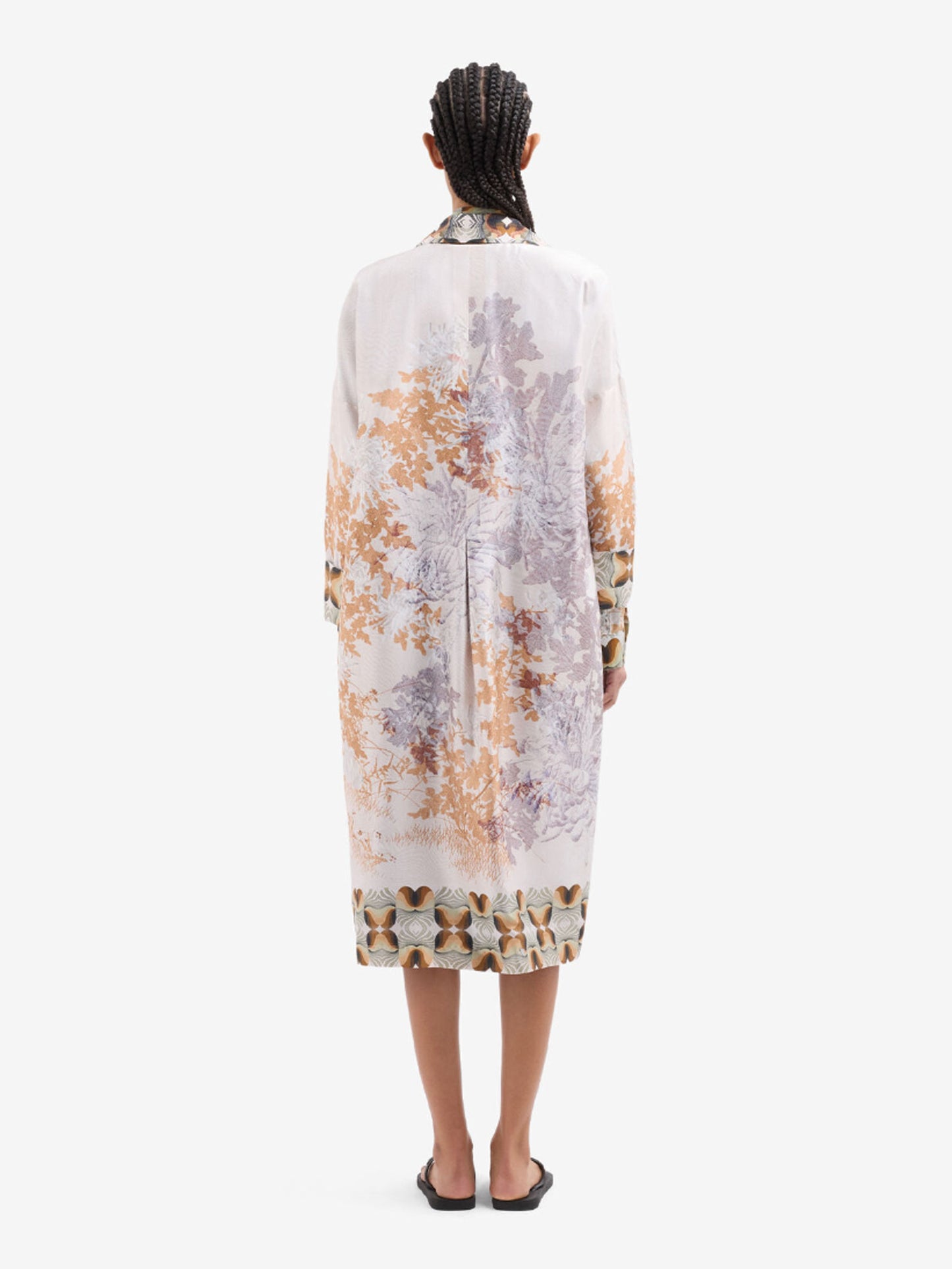 A model viewed from the back wearing a long jacket with a muted botanical pattern in beige, brown, and gray, paired with black slide sandals against a plain white background.