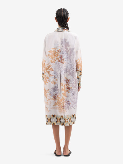 A model viewed from the back wearing a long jacket with a muted botanical pattern in beige, brown, and gray, paired with black slide sandals against a plain white background.