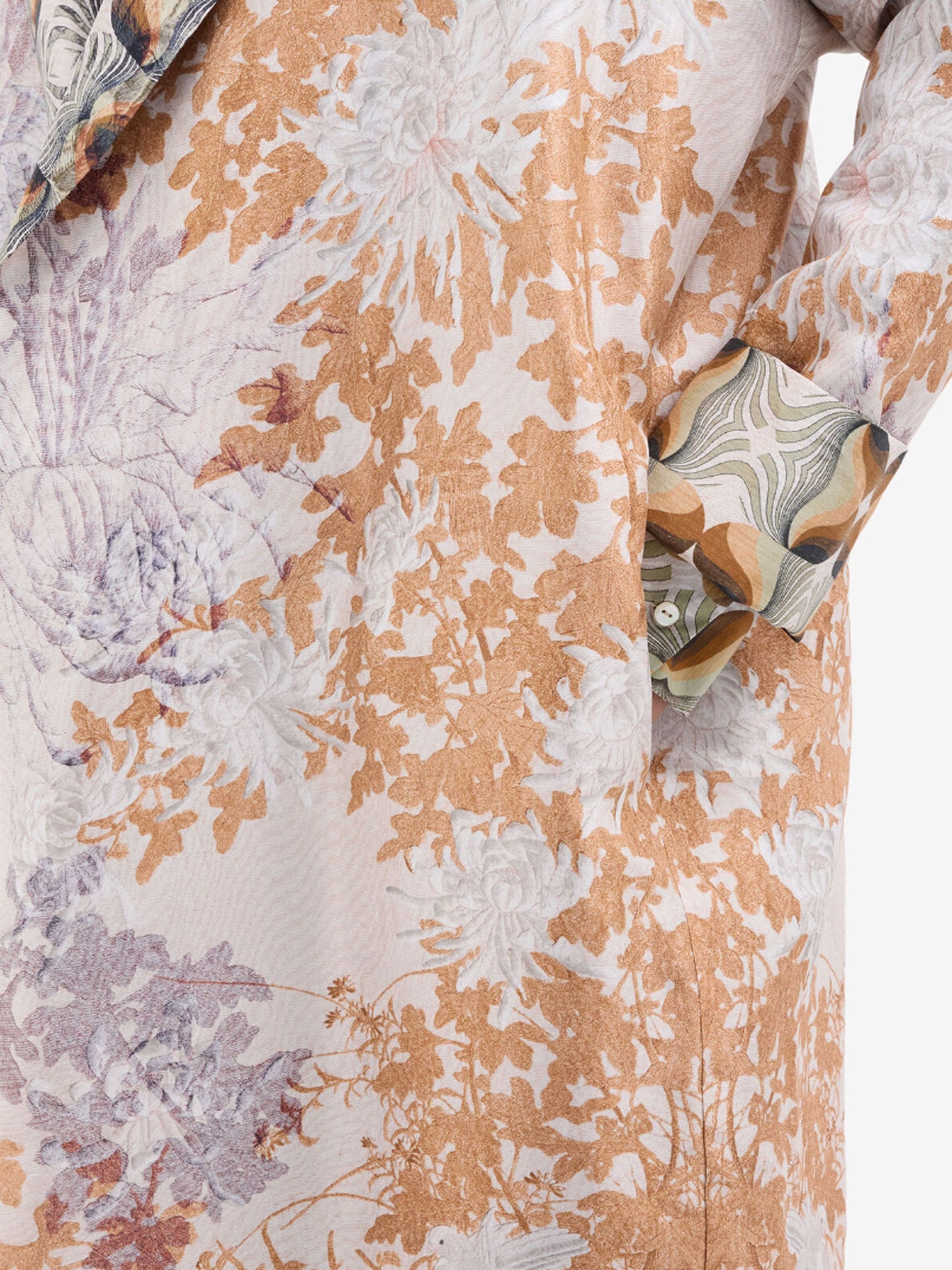 Close-up of a patterned fabric garment featuring a floral and botanical design in warm tan, beige, and muted lavender-gray tones on an off-white base, with a matching patterned pocket flap.