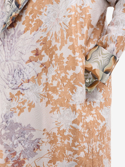 Close-up of a patterned fabric garment featuring a floral and botanical design in warm tan, beige, and muted lavender-gray tones on an off-white base, with a matching patterned pocket flap.