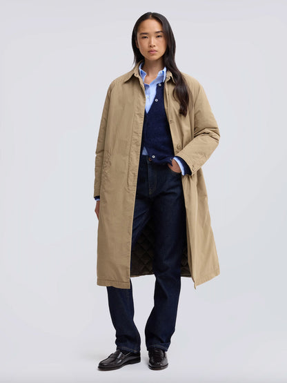 A model stands against a light gray studio background wearing a long beige trench coat over a light blue button-up shirt, navy sweater vest, navy jeans, and black shoes.