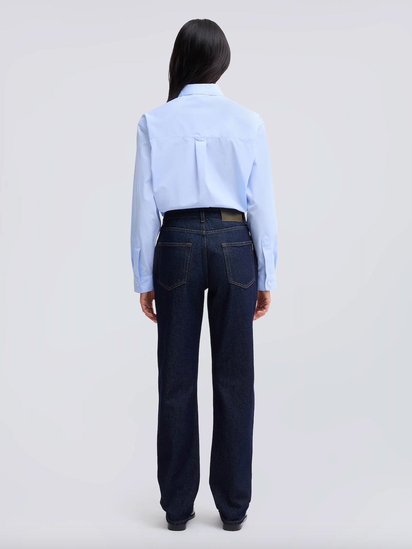 A model viewed from the back wearing a light blue long-sleeve button-up shirt tucked into dark blue straight-leg jeans, set against a plain light gray studio background.