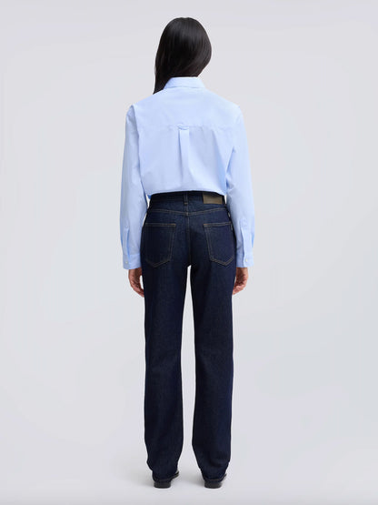 A model viewed from the back wearing a light blue long-sleeve button-up shirt tucked into dark blue straight-leg jeans, set against a plain light gray studio background.