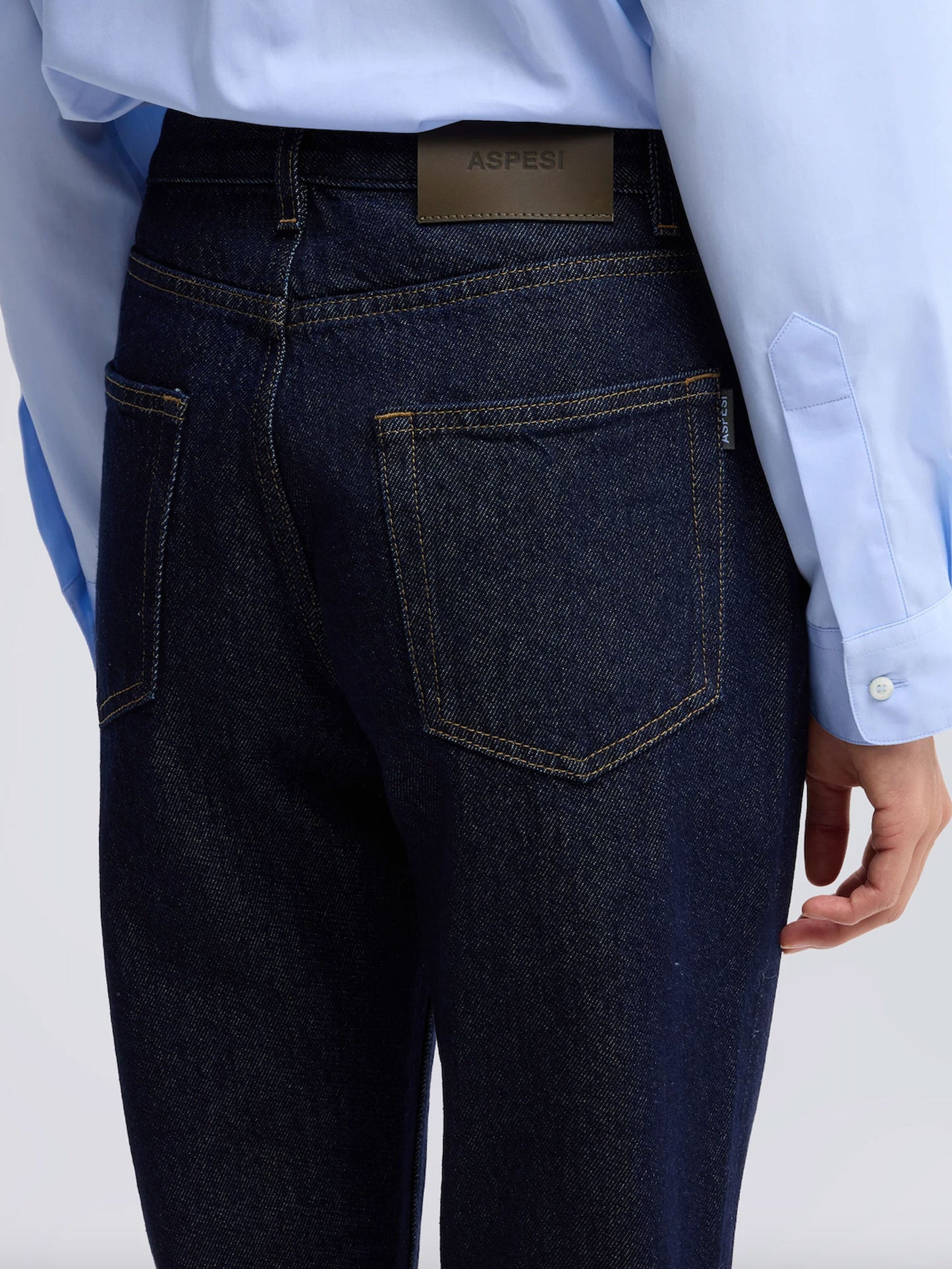 Close-up back view of a person wearing dark indigo denim jeans with a light blue dress shirt tucked in. A tan leather ASHRI brand patch is visible at the back waistband.