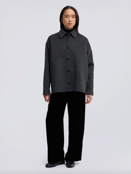 A model stands against a light gray studio background wearing a dark charcoal gray button-front collared jacket and black wide-leg trousers.