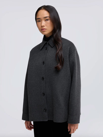 A woman modeling a dark gray oversized button-up coat with a pointed collar over a black turtleneck, set against a plain light gray studio background.