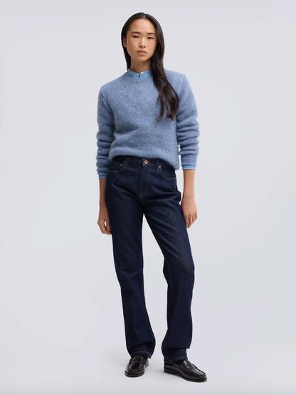 A model stands against a light gray studio background wearing a light blue knit mock-neck sweater, dark blue jeans, and black leather shoes.