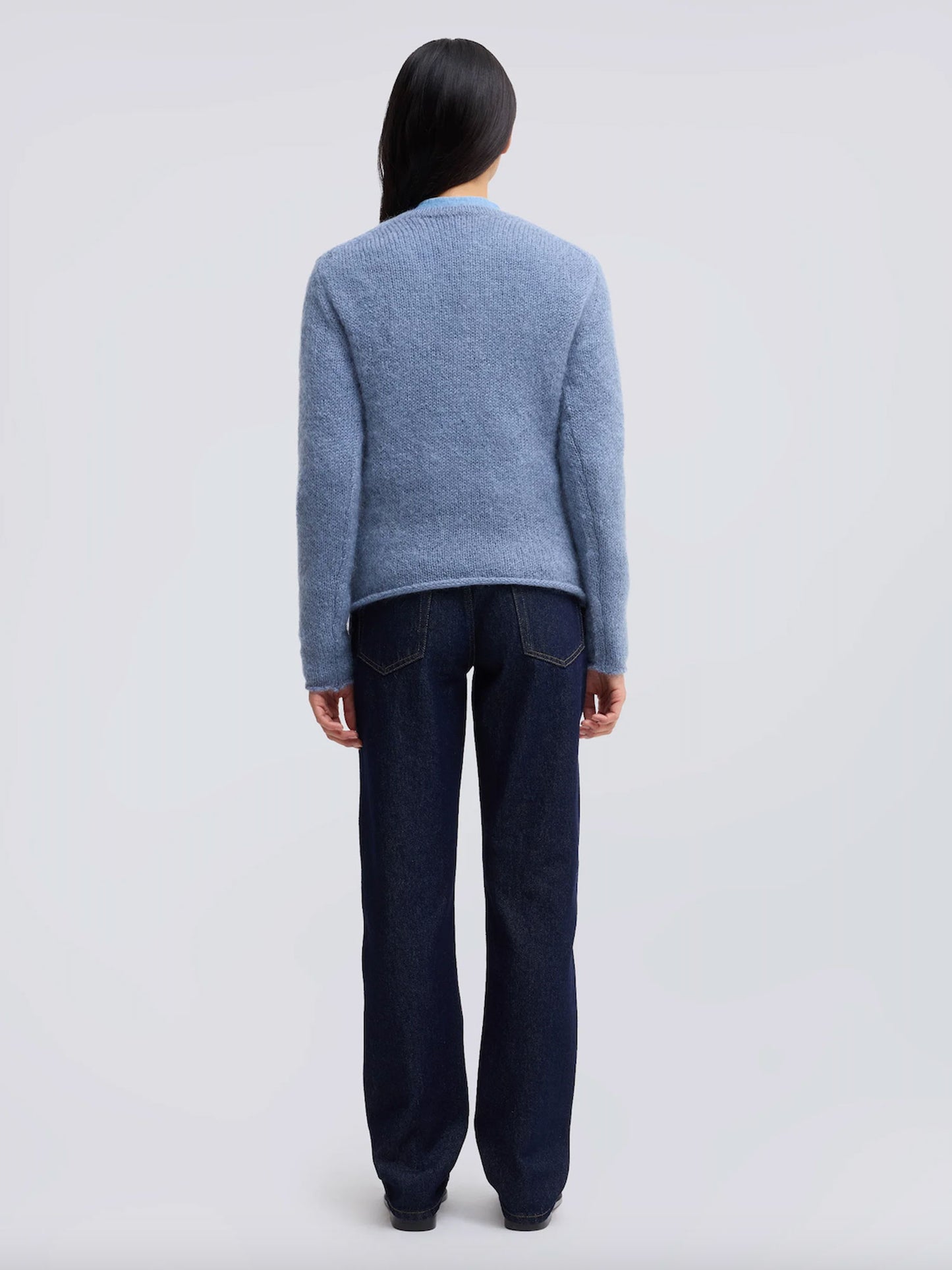 A model viewed from the back wearing a light blue knit sweater and dark blue jeans against a light gray studio background.