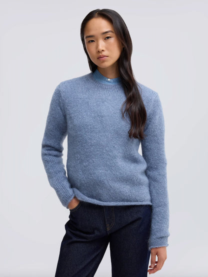 A woman modeling a light blue textured knit sweater with a mock neck, paired with dark blue jeans, against a plain light gray studio background.