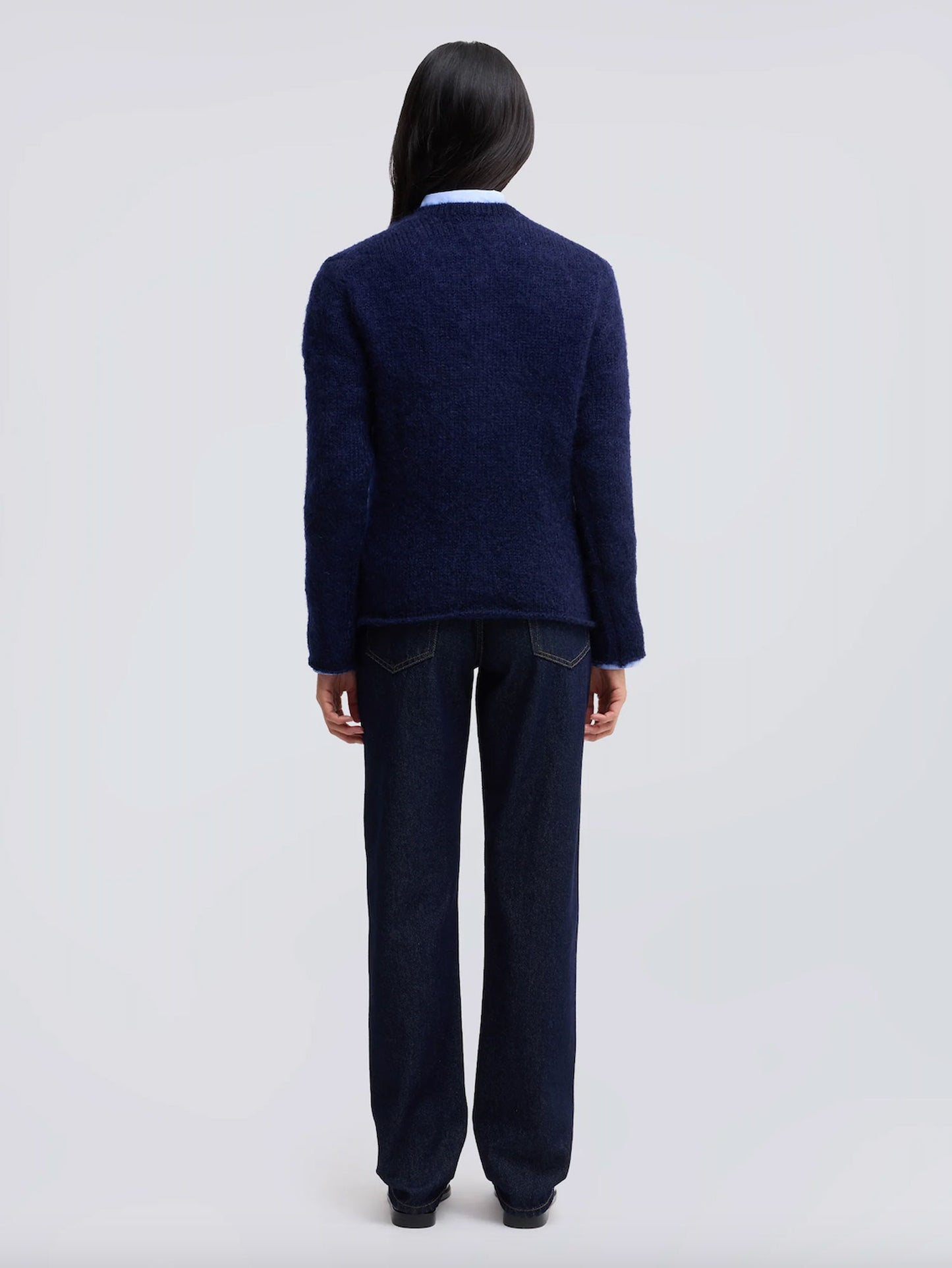 A model viewed from the back wearing a dark navy blue textured knit cardigan over a light blue collared shirt and dark navy wide-leg jeans, set against a plain light gray studio background.