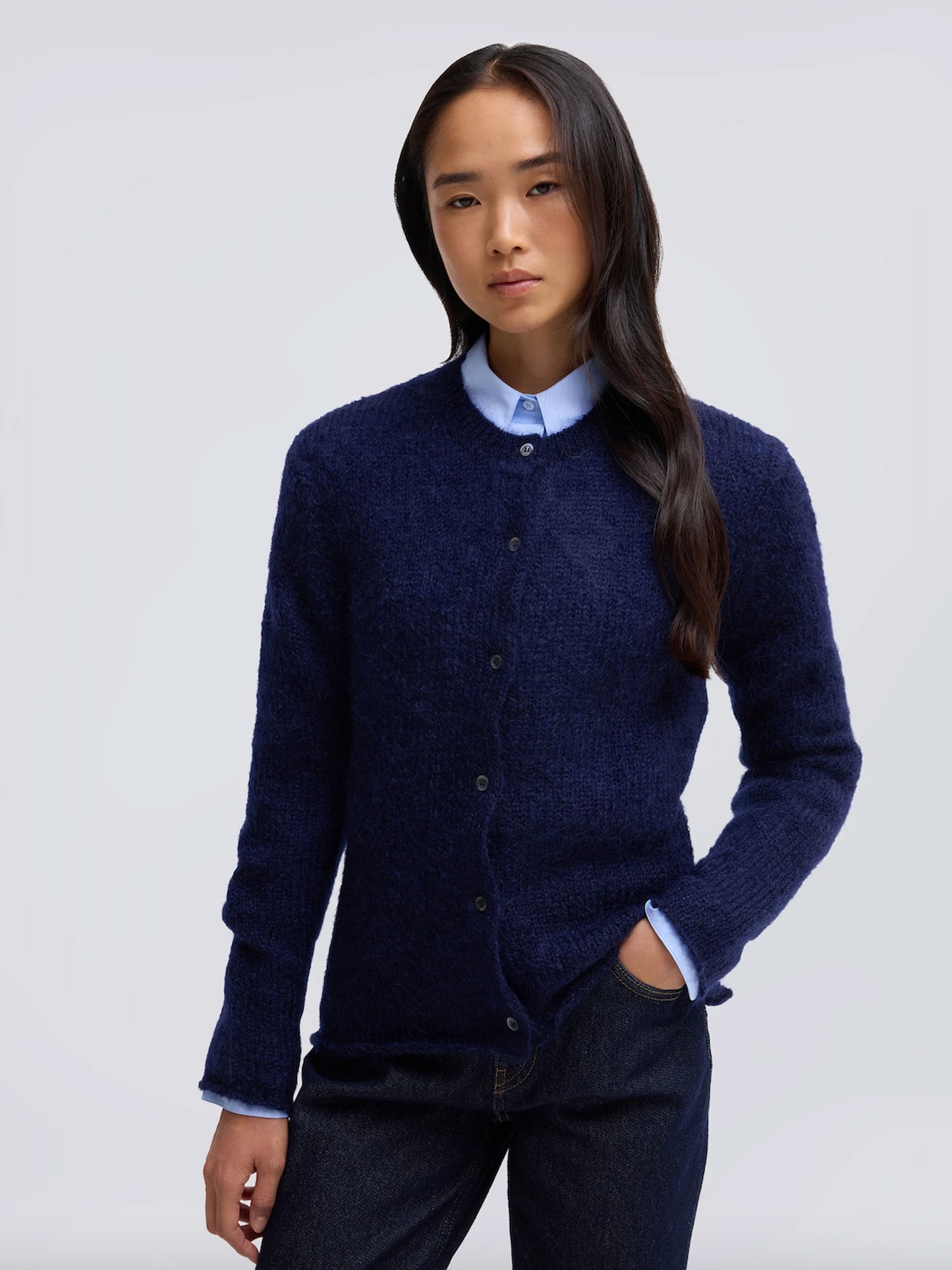 A woman modeling a dark navy blue textured knit cardigan sweater over a light blue collared shirt, paired with dark jeans, against a plain light gray studio background.