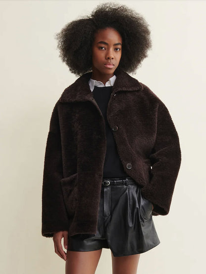 A model with an afro hairstyle wearing a dark brown plush teddy coat over a black top and black leather shorts, photographed against a plain light beige background.