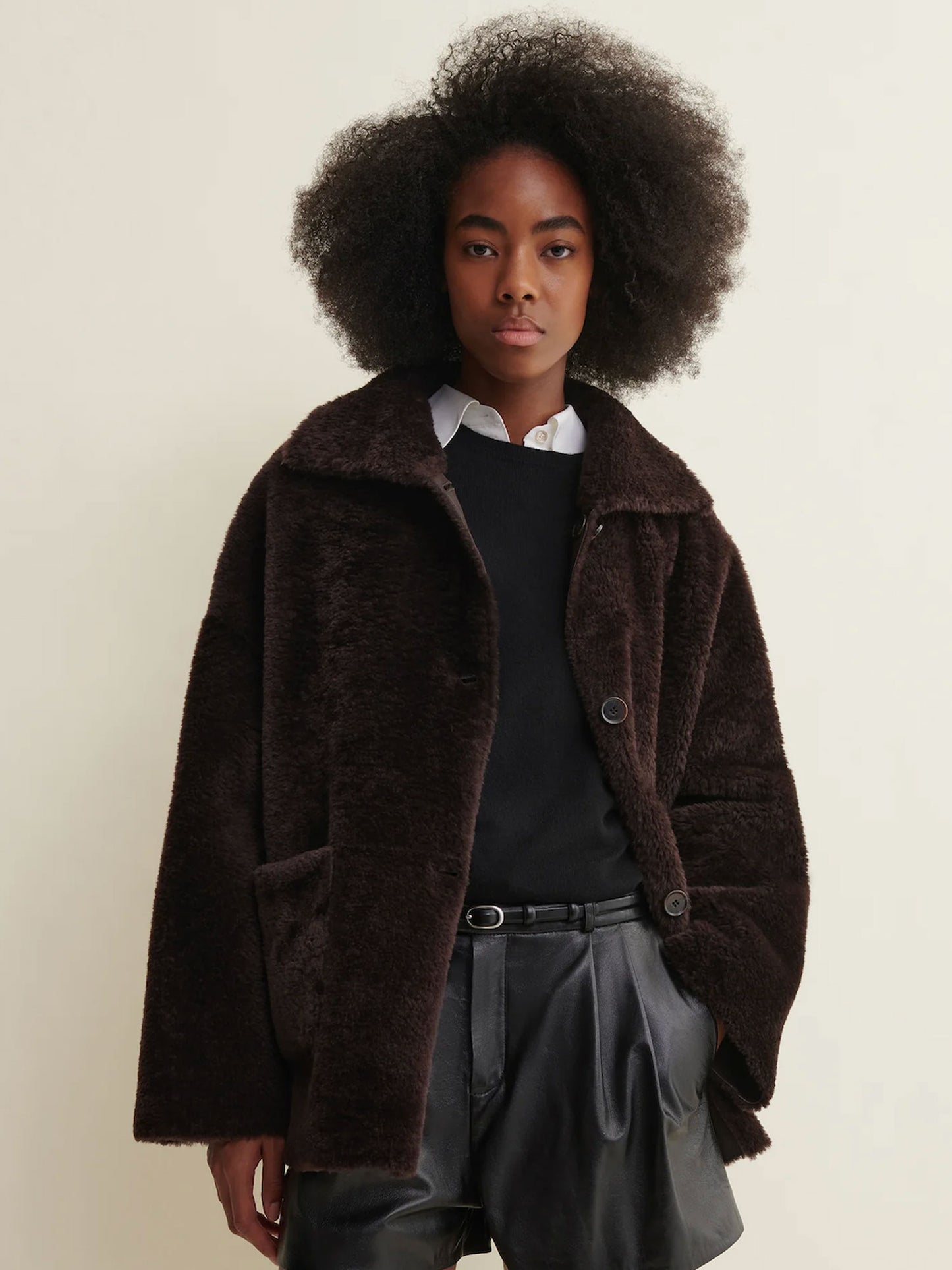 A model with an afro hairstyle wearing a dark brown fuzzy teddy coat over a black top and gray pants, standing against a plain off-white background.