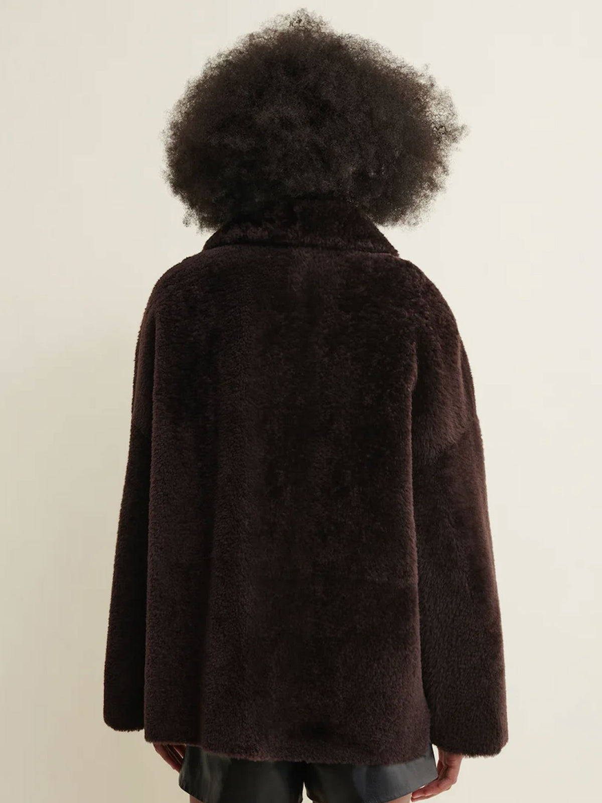 A person shown from the back wearing a dark brown fuzzy teddy-fur style jacket against a plain light beige studio background.
