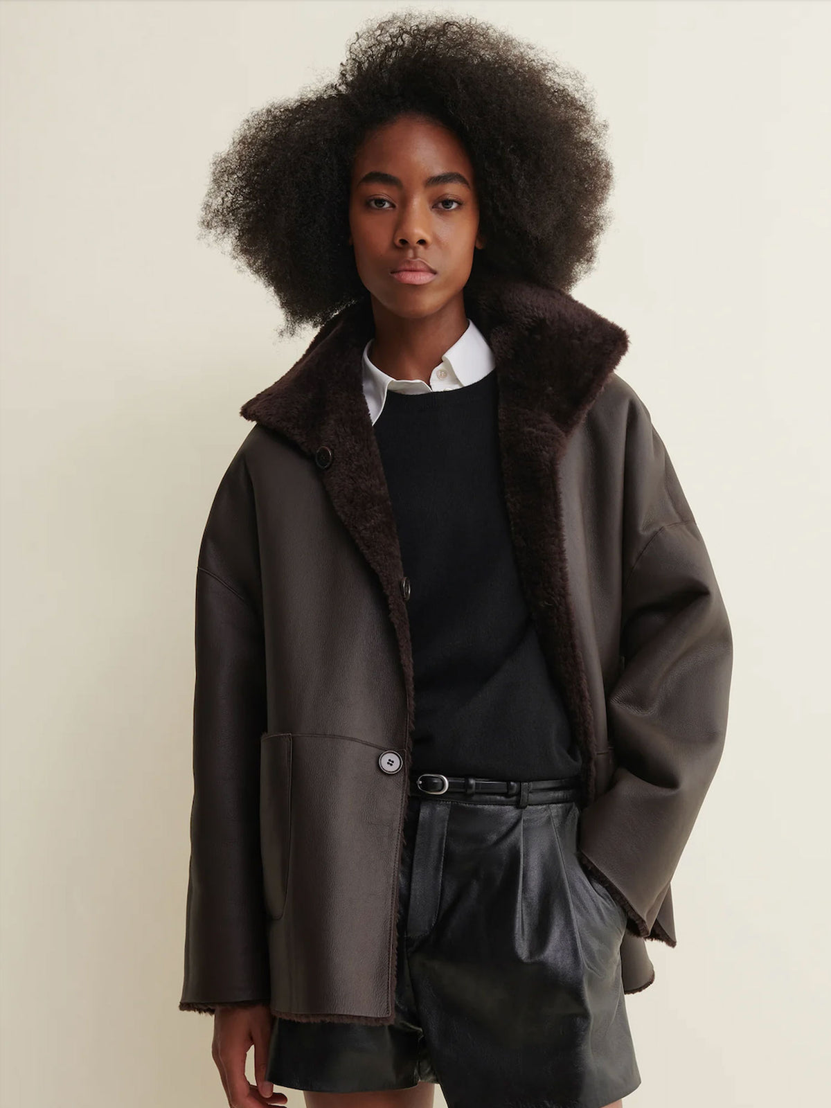 A model with an afro hairstyle wearing a dark brown shearling-lined jacket over a black sweater and white collared shirt, paired with black leather shorts, against a plain beige background.
