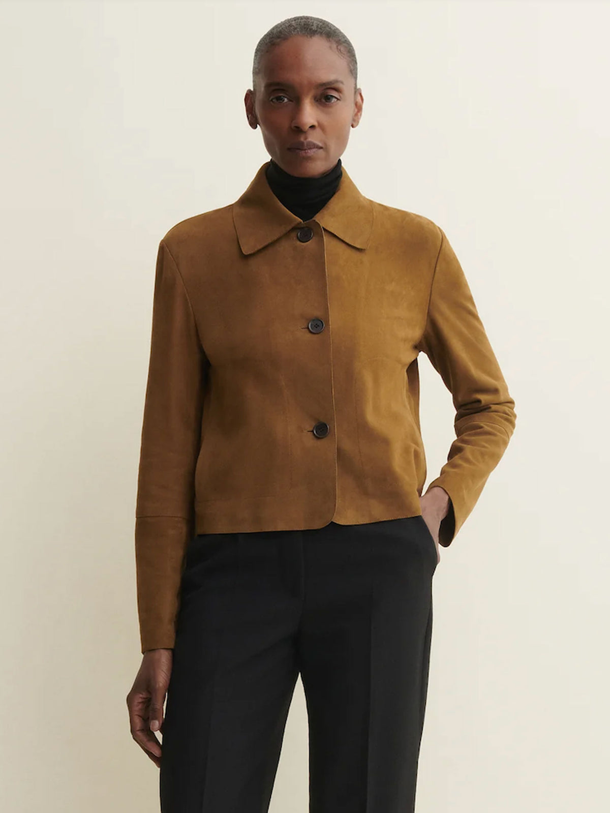 A model wearing a cropped, long-sleeve suede jacket in a warm camel brown color, styled over a black turtleneck and black trousers, against a plain off-white background.