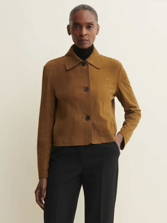 A model wearing a cropped, long-sleeve suede jacket in a warm camel brown color, styled over a black turtleneck and black trousers, against a plain off-white background.