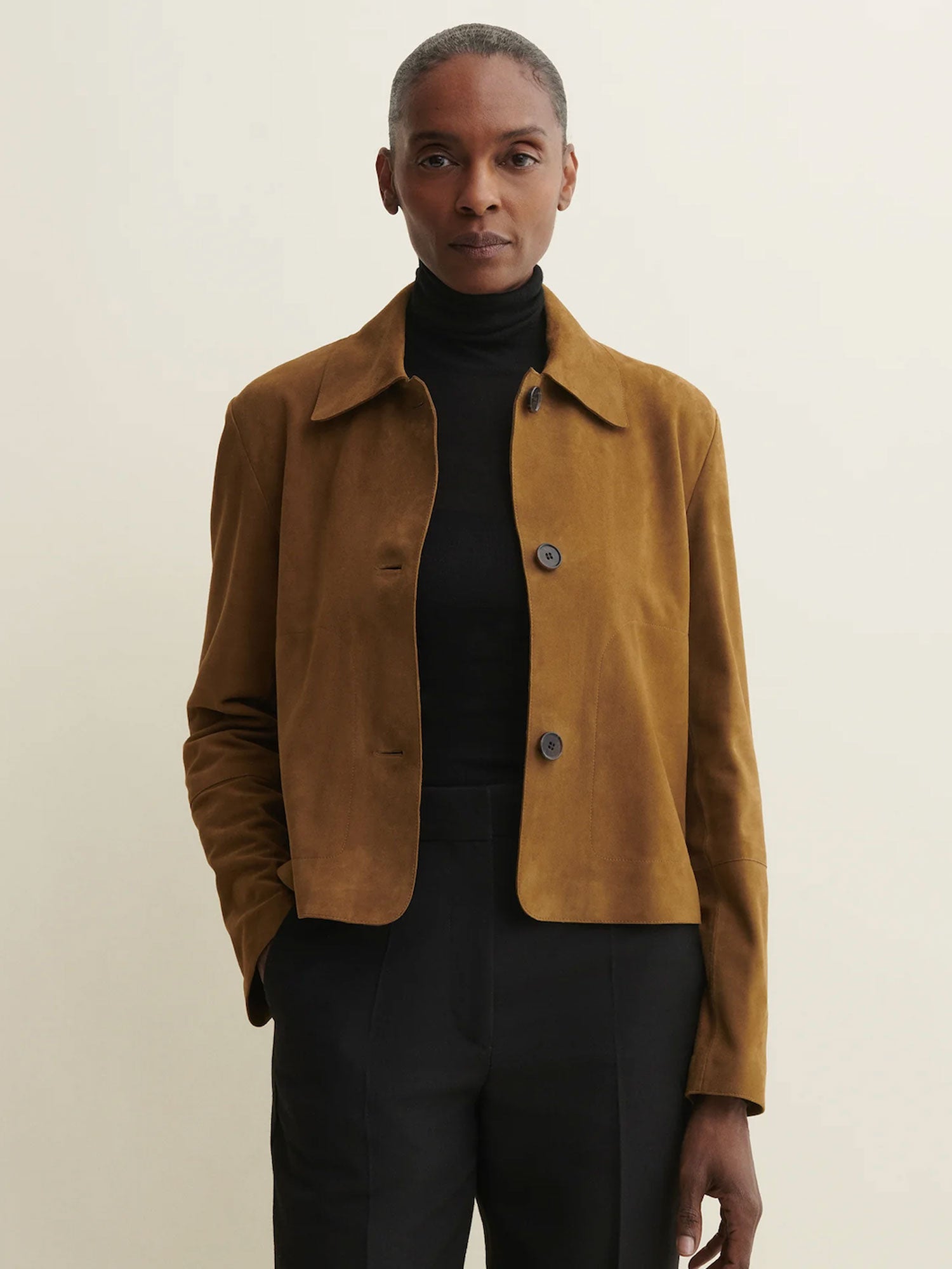 A model wearing a cropped tan suede jacket over a black turtleneck and black trousers, standing against a plain light beige background.