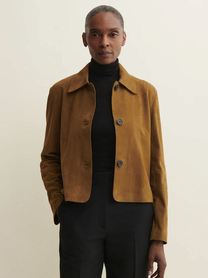 A model wearing a cropped tan suede jacket over a black turtleneck and black trousers, standing against a plain light beige background.