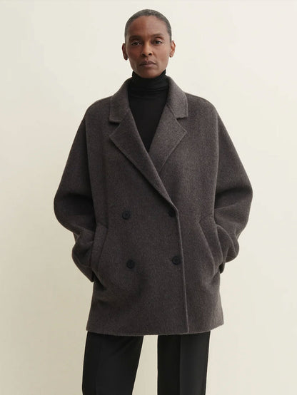 A model wearing a dark gray double-breasted wool-blend coat over a black turtleneck and black trousers, standing against a plain light beige background.