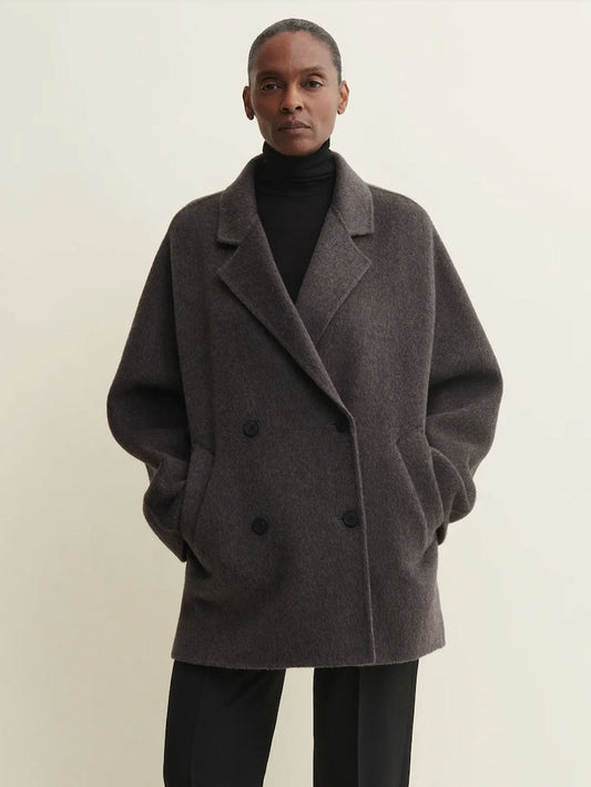 A model wearing a dark gray double-breasted wool-blend coat over a black turtleneck and black trousers, standing against a plain light beige background.