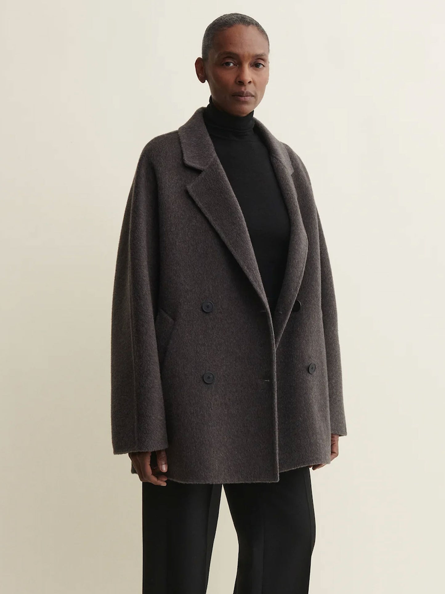 A model wearing an oversized, double-breasted dark brown wool-blend coat over a black turtleneck and black trousers, standing against a plain off-white studio background.