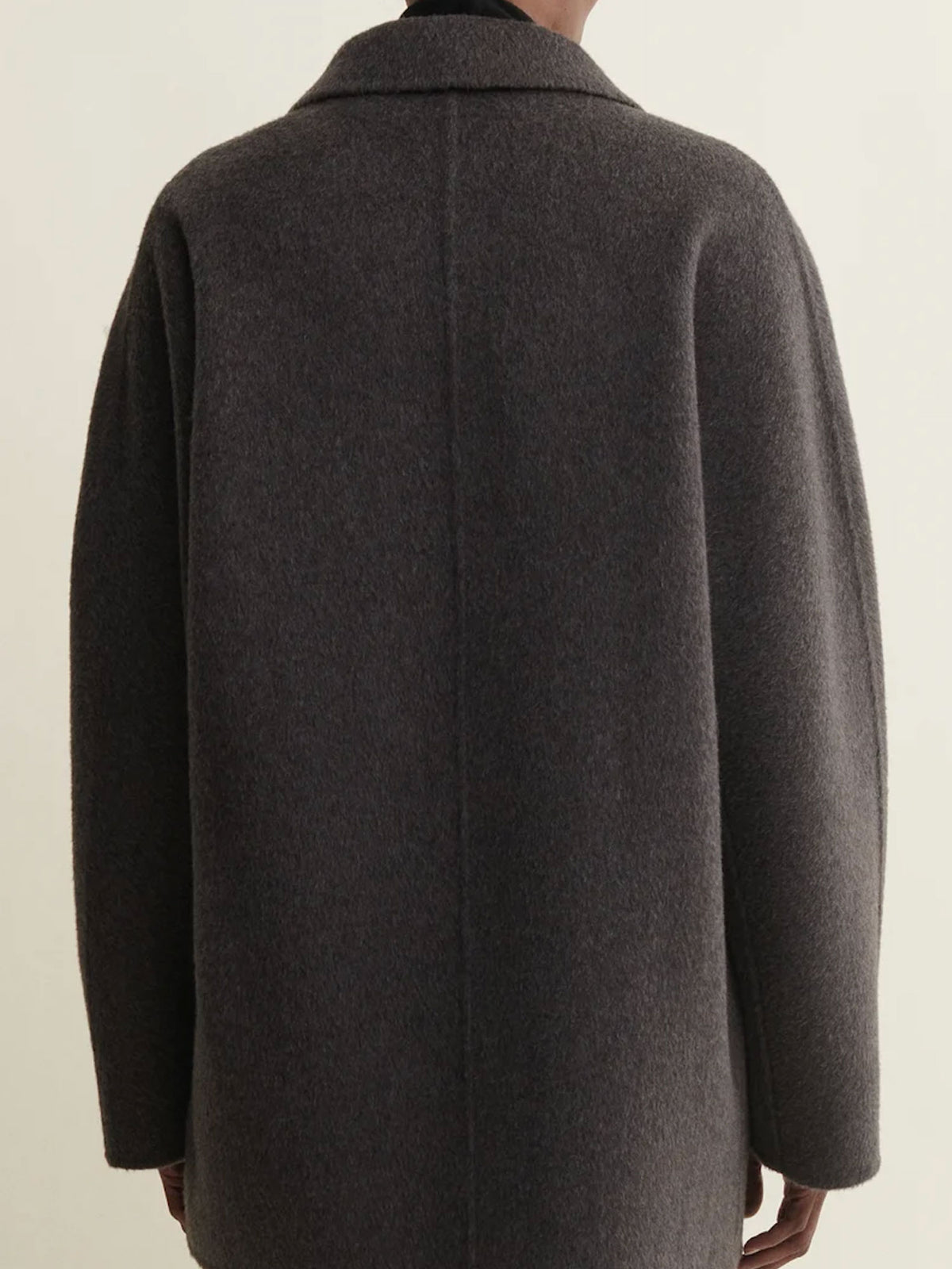 A dark charcoal gray wool coat shown from the back on a model, featuring a wide collar and long sleeves, against a plain off-white background.