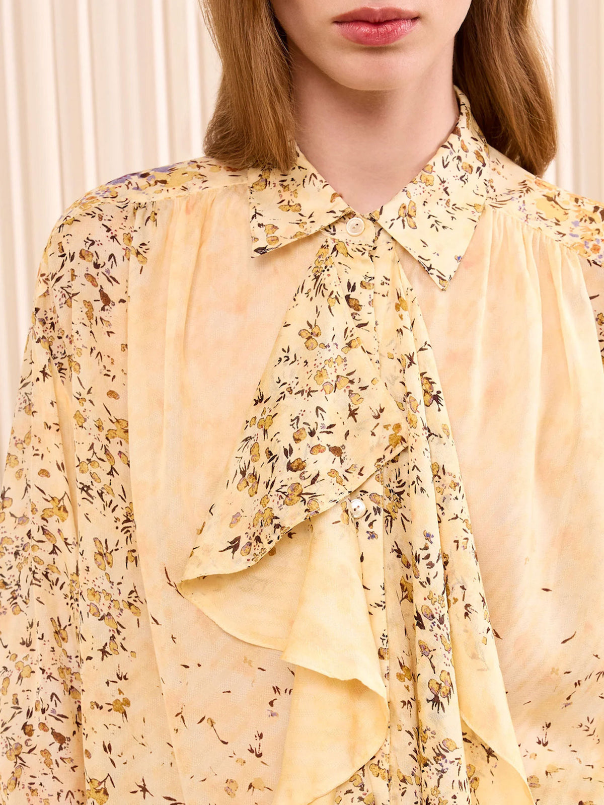 A close-up of a model wearing a pale yellow blouse with a delicate brown and black floral print, featuring a ruffled tie-neck detail and a pointed collar.