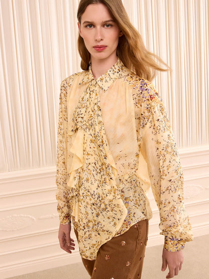 A woman wearing a long-sleeve ruffled blouse with a yellow and beige floral print, paired with brown pants, standing against a cream-colored paneled wall.