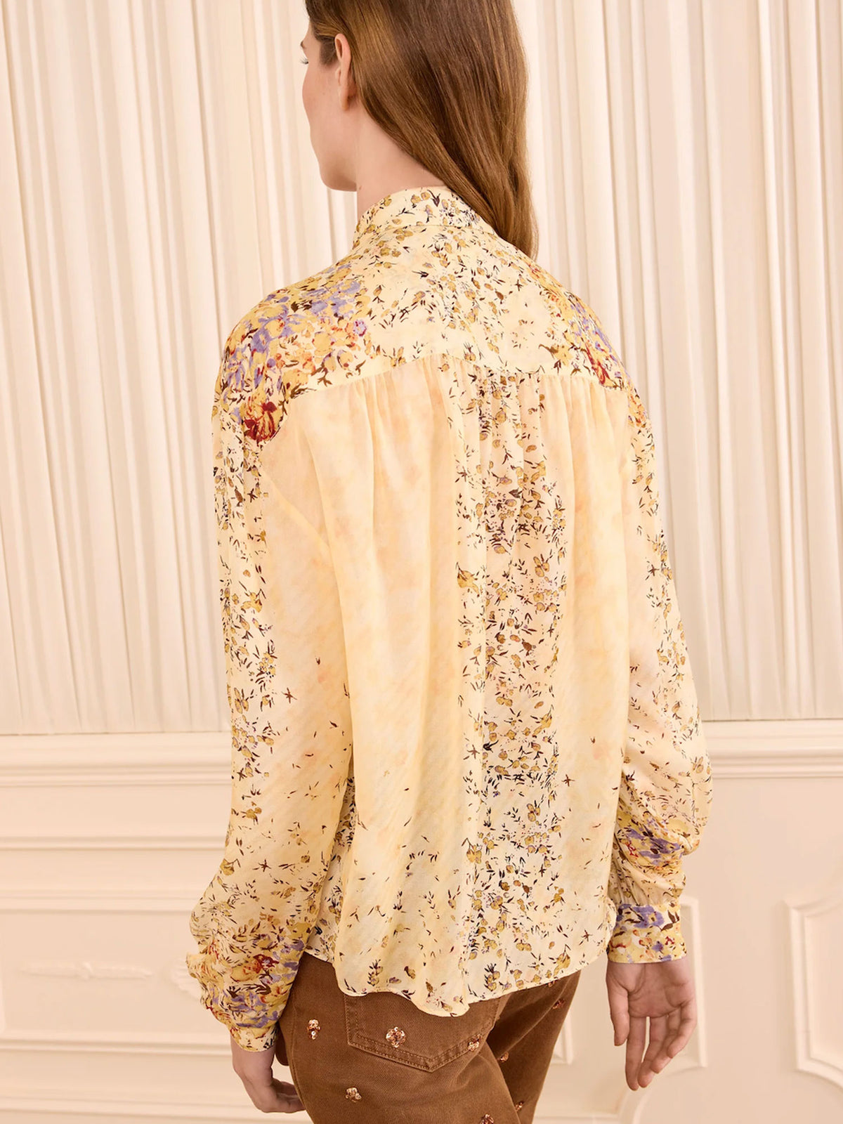 A woman shown from the back wearing a long-sleeve cream blouse with a delicate multicolor floral print and gathered shoulder details, paired with brown pants, set against a cream paneled wall.